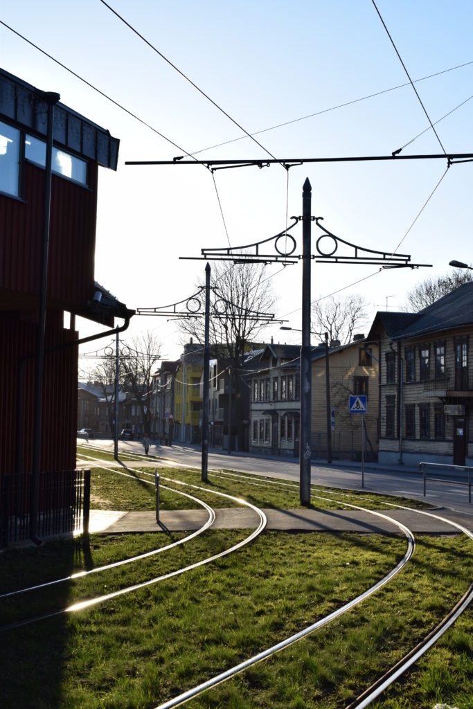 Keskellä kaksi ratikkakiskoa kulkevat rinnakkain, vasemmalla rakennus, oikealla asuintaloja. Aurinko paistaa ja ruoho vihertää.