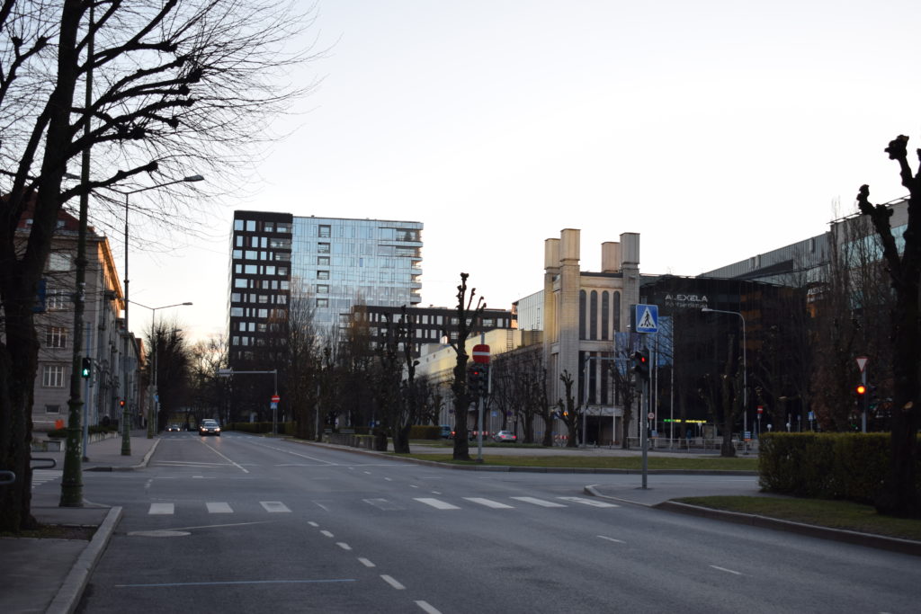 Edessä kaupunkimaisemaa: lasinen kerrostalo, historiallisen näköinen matalampi rakennus. Tyhjät tiet, ilta-aurinko paistaa.