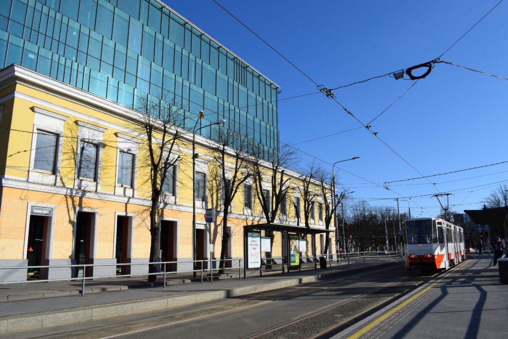 Keskekllä tyhjä katu ja tyhjä ratikkalaituri, johon saapuu juuri ratikka. Vasemmalla keltainen rakennus ja lasirakennus. Aurinko paistaa, kadut ovat tyhjät.