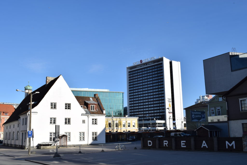 Vasemmalla valkoinen matala tiilitalo, johon aurinko paistaa kirkkaasti. Horisontissa Viru-hotelli ja muita rakennuksia. Oikealla edessä kirjaimet D R E A M kiinnitetty aitaan. 